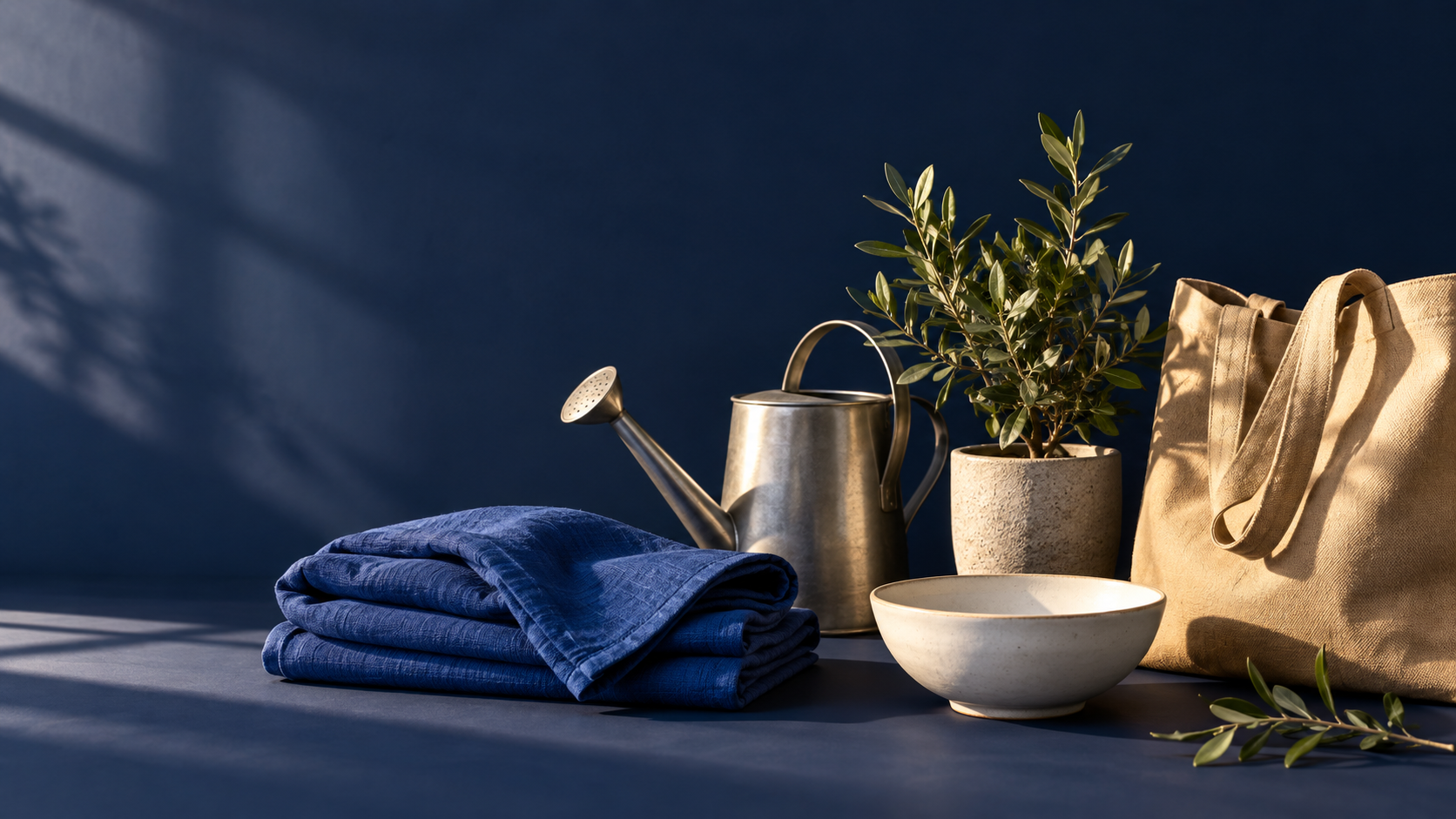 Composition d’univers : textile bleu, arrosoir en métal, plante, bol en céramique et sac en toile sur fond bleu nuit.